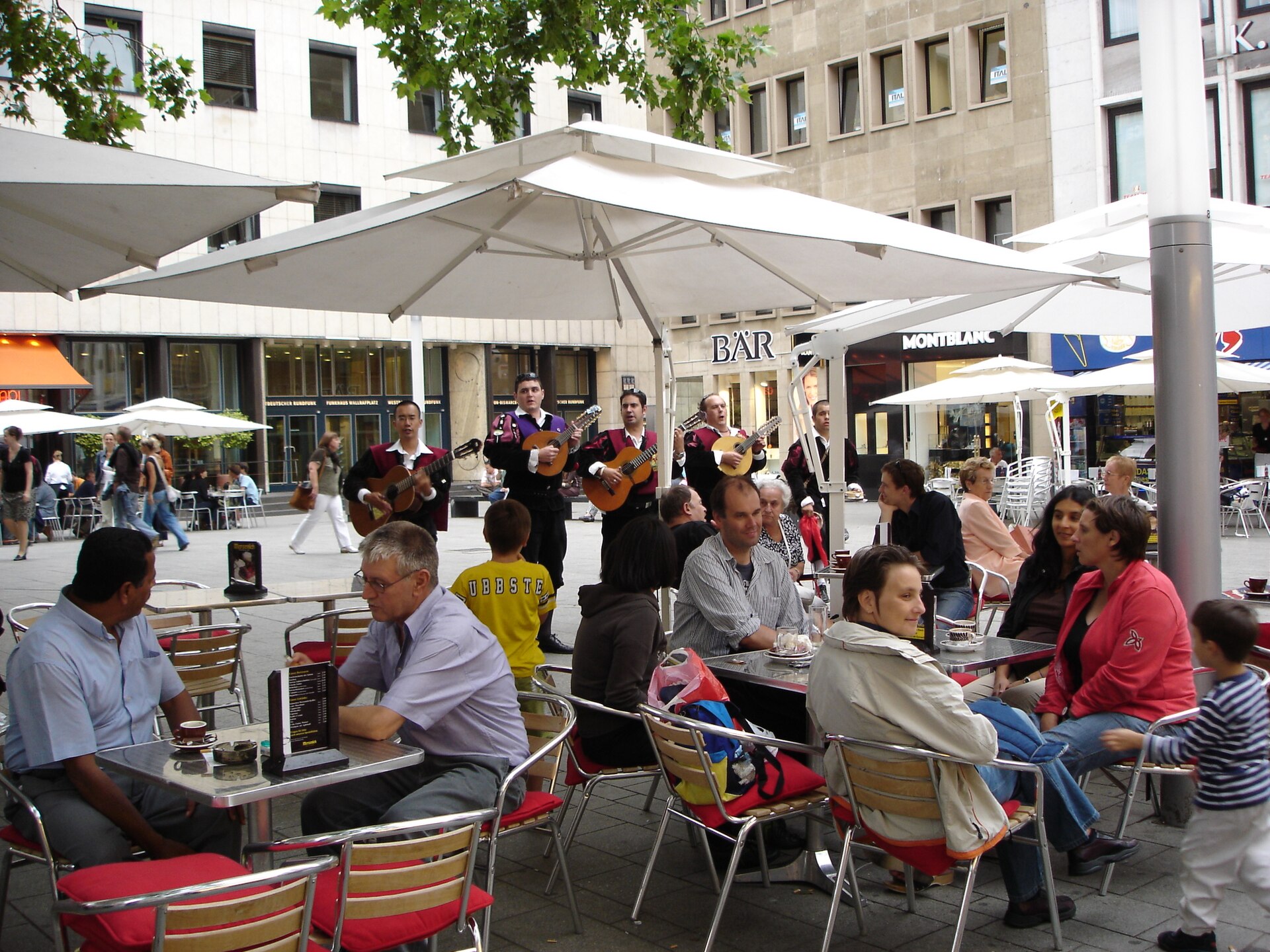 Musiker spielen auf einem Platz in einem Kölner Veedel zwischen Café-Tischen und Sonnenschirmen – ein möglicher Spielort der Fête, schwarz-weiß